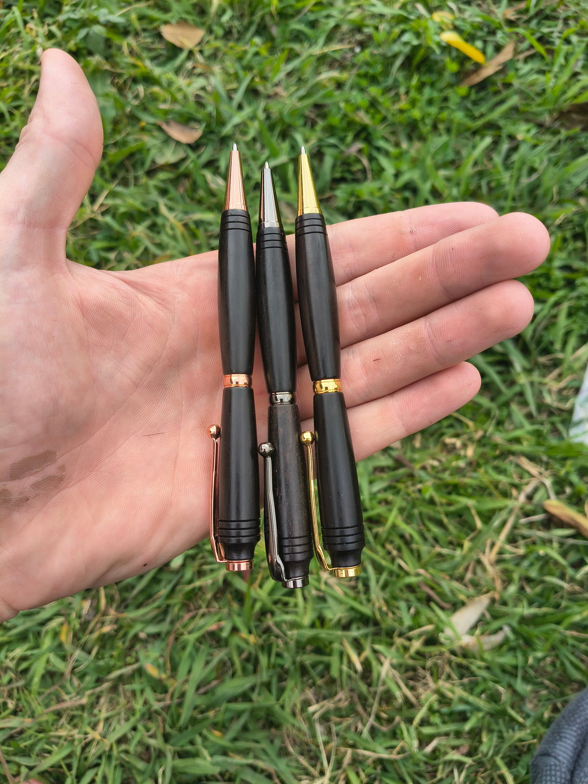 Four black ballpoint pens with gold accents are held in an open hand over a green grass background.