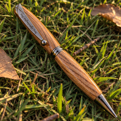 A wooden ballpoint pen with a natural wood grain handle and silver metal accents, resting on green grass with dry leaves.