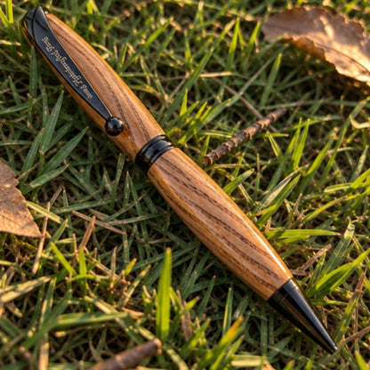 A wooden ballpoint pen with a black clip and tip lies diagonally on green grass with dry leaves, featuring the engraved brand name 'Redhead Handcrafted Pens'.
