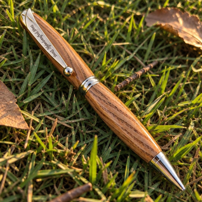 A wooden ballpoint pen with a polished wood grain handle and silver metal accents, resting diagonally on green grass with a few dry leaves.