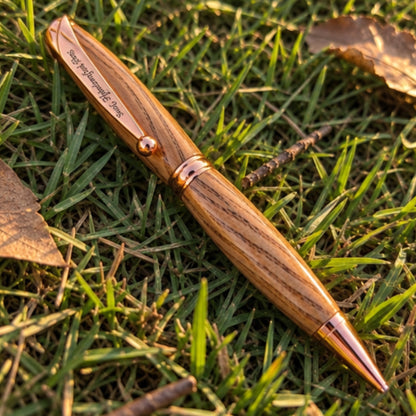 A wooden ballpoint pen with a polished oak wood grain finish and rose gold metal accents, resting diagonally on green grass with dry leaves.