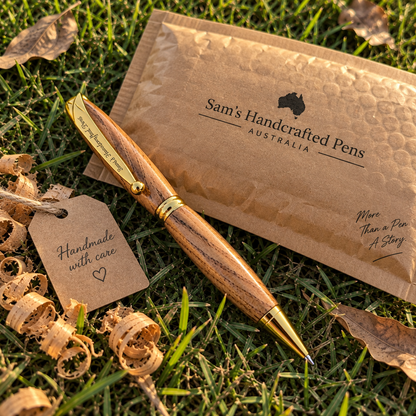 A wooden pen with gold metal accents lies on green grass beside a brown kraft paper pouch labeled 'Sam's Handcrafted Pens Australia' and a small tag reading 'Handmade with care.'.