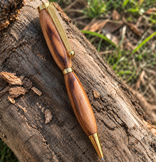 A wooden ballpoint pen with a polished brown wood body and gold metal accents, resting diagonally on a textured tree log outdoors with grass and soil in the background.