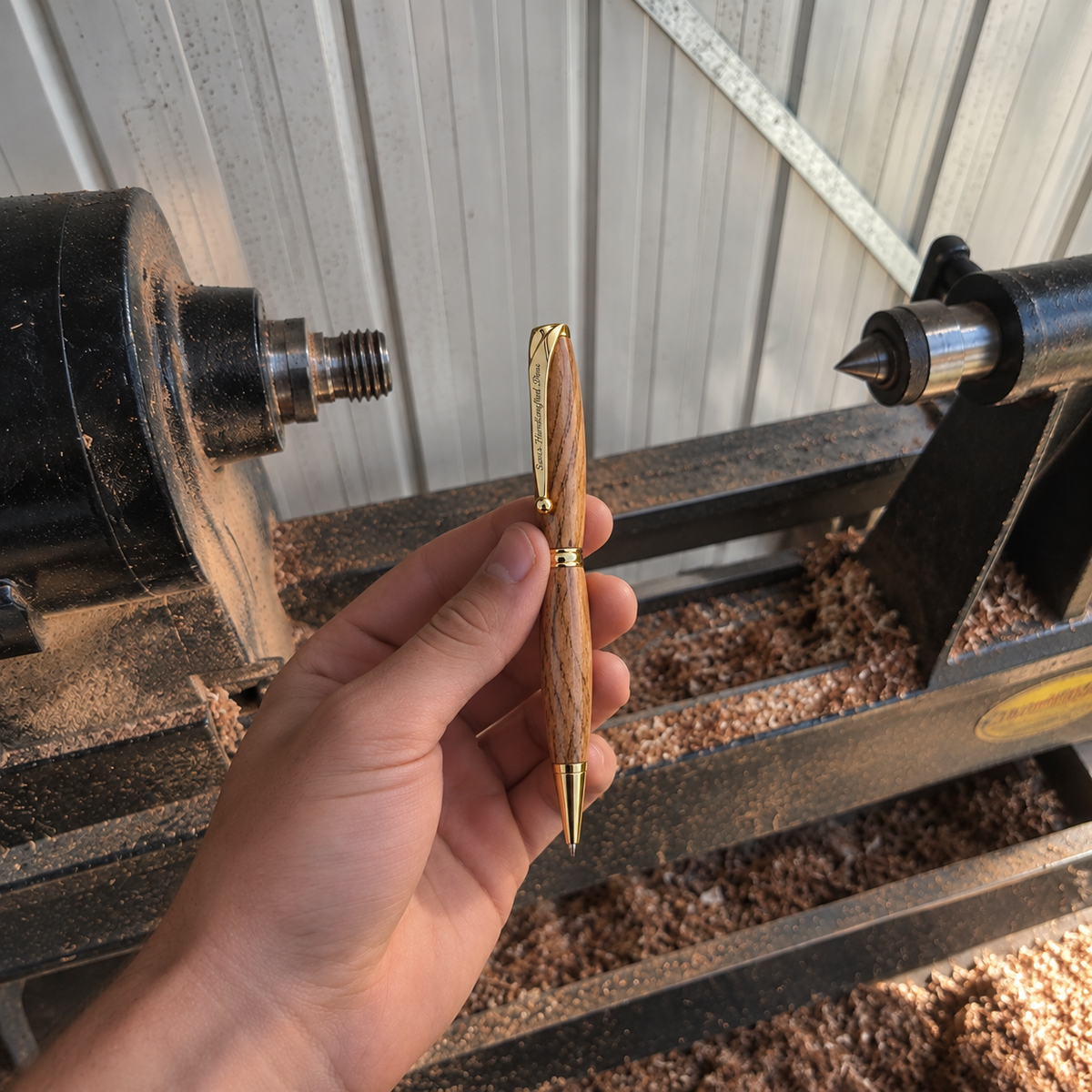 A hand holds a slim pen with a natural wood grain body and gold metal accents, positioned in front of a woodworking machine with sawdust in the background.