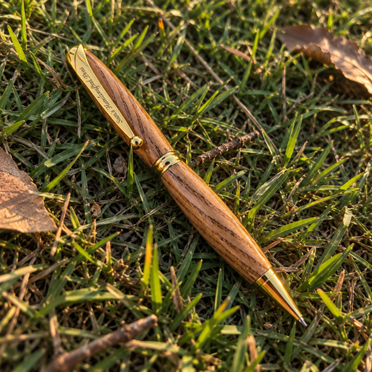 A wooden pen with a gold-plated clip and trim lies diagonally on a patch of green grass with scattered dry leaves.