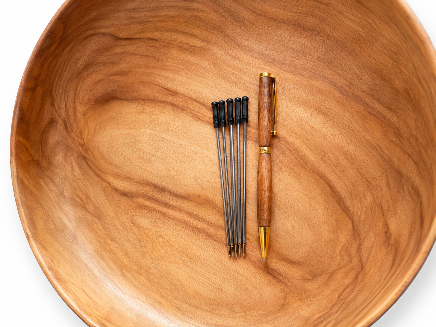 A wooden bowl with a rich grain pattern serves as a background for a pen and a set of arrow shafts. The pen features a warm brown wooden handle, gold-toned metal accents, and a gold clip.