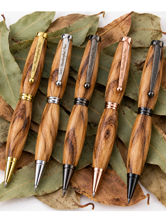 Five wooden ballpoint pens with different metal clip and tip finishes are arranged on a bed of dried green and brown leaves. The pens feature natural wood grain bodies with gold, silver, rose gold, and black accents.
